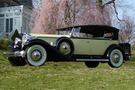 The 1932 Packard Phaeton Model 905 sits in front of a blooming cherry tree. The car is a light green/yellow with Black highlights, features white wheels, and has an extra wheel attached to the side of the car.
