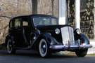 The 1937 Packard Model 1508 sits in front of a white arch. It is a fully black car.