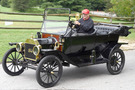 An older gentleman with a red baseball cap is driving the 1914 Ford Model T on a paved road in front of a field. The car is mostly black save for a red panel near the front of the car