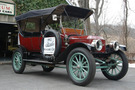 The 1912 Stanley model 87 has a firetruck red body and mint green wheel spokes and under carriage. The top of the car is up, covering the seats from above while leaving the sides of the car open 