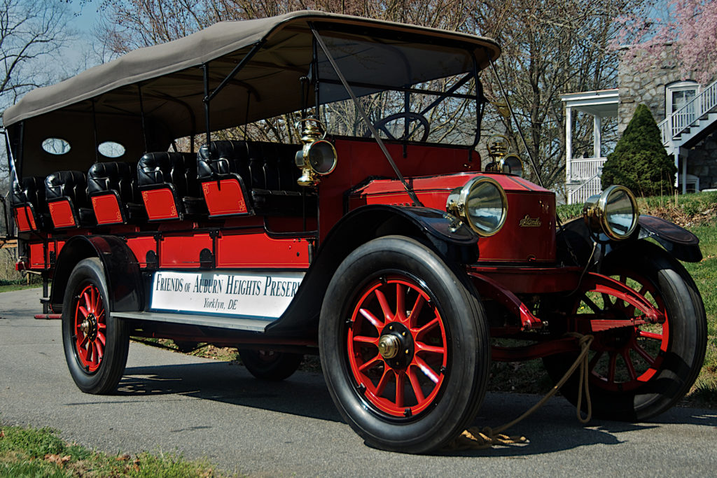 The 1915 Stanley Model 820 Mountain Wagon has five rows of seats, fitting across approximately three people per row. The car is all red with a black top. There is a sign on the side that reads "Friends of Auburn Heights Preserve, Yorklyn, DE"